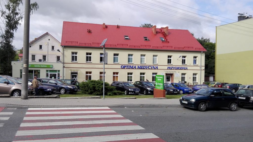 Budynek przychodni lekarskiej z apteką w Prudniku, przed nim parking pełen samochodów.