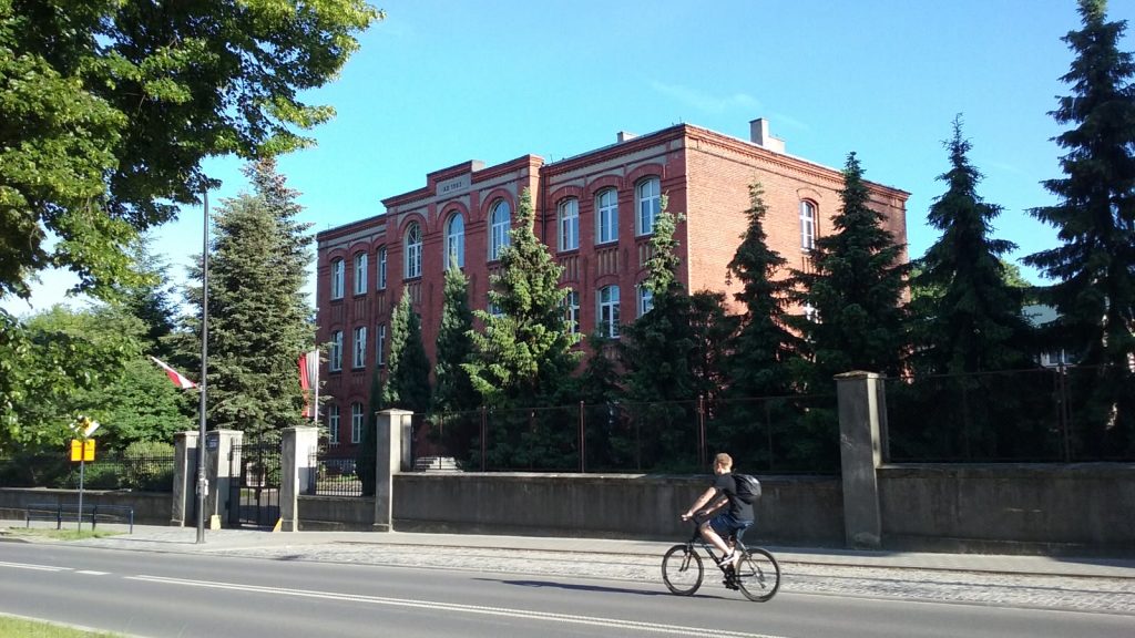 Ceglany, trzykondygnacyjny budynek liceum ogólnokształcącego przy ulicy, otoczony świerkami, w słoneczny dzień.
