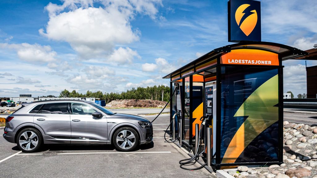 Srebrny elektryczny SUV podłączony kablem do szybkiej ładowarki na przydrożnej stacji ładowania w Norwegii, w tle błękitne niebo i las.