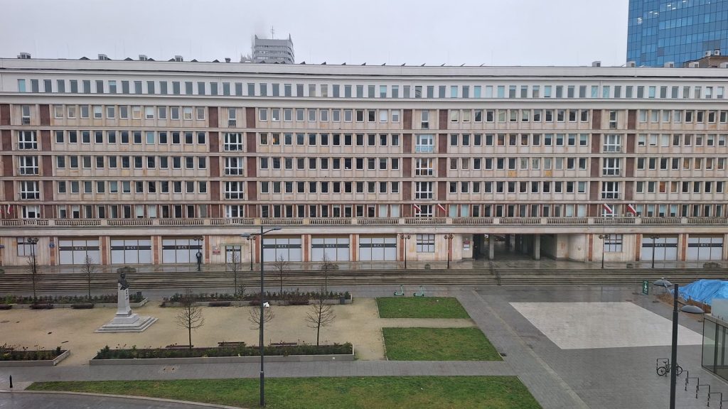 Plac Powstańców Warszawy w pochmurny zimowy dzień: przed długim, jasnym budynkiem widać podniesione rabaty z młodymi, bezlistnymi drzewami, pas trawnika i pomnik.