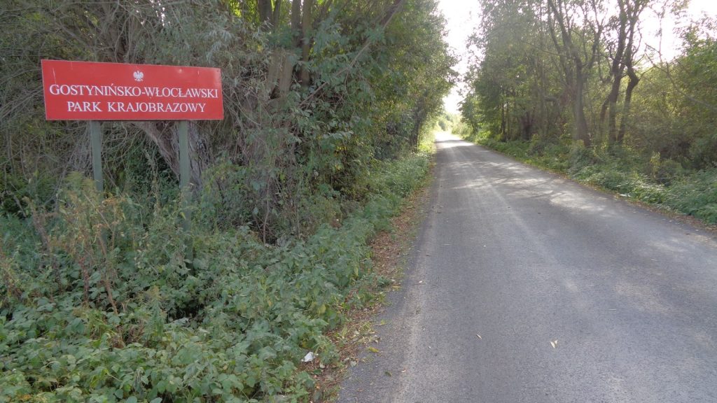 Czerwona tablica z napisem Gostynińsko-Włocławski Park Krajobrazowy stojąca przy wąskiej drodze wśród drzew.