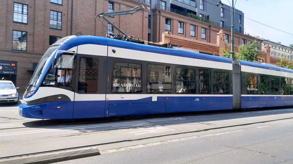 Niebiesko-biały, wieloczłonowy tramwaj Krakowiak jadący ulicą w Krakowie wzdłuż ceglanych kamienic.