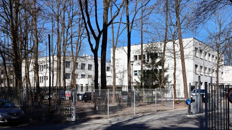 Białe budynki Narodowego Instytutu Kardiologii w warszawskim Aninie, widziane zza ogrodzenia spomiędzy bezlistnych drzew.