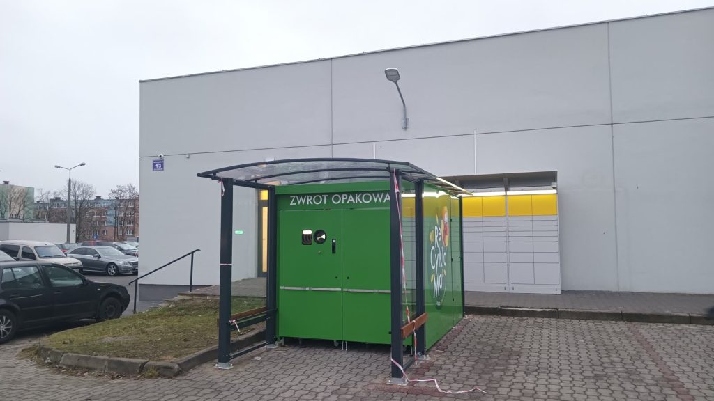 Zielony automat do zwrotu opakowań z napisem „Zwrot opakowań” pod wiatą przed ścianą supermarketu na osiedlowym parkingu.
